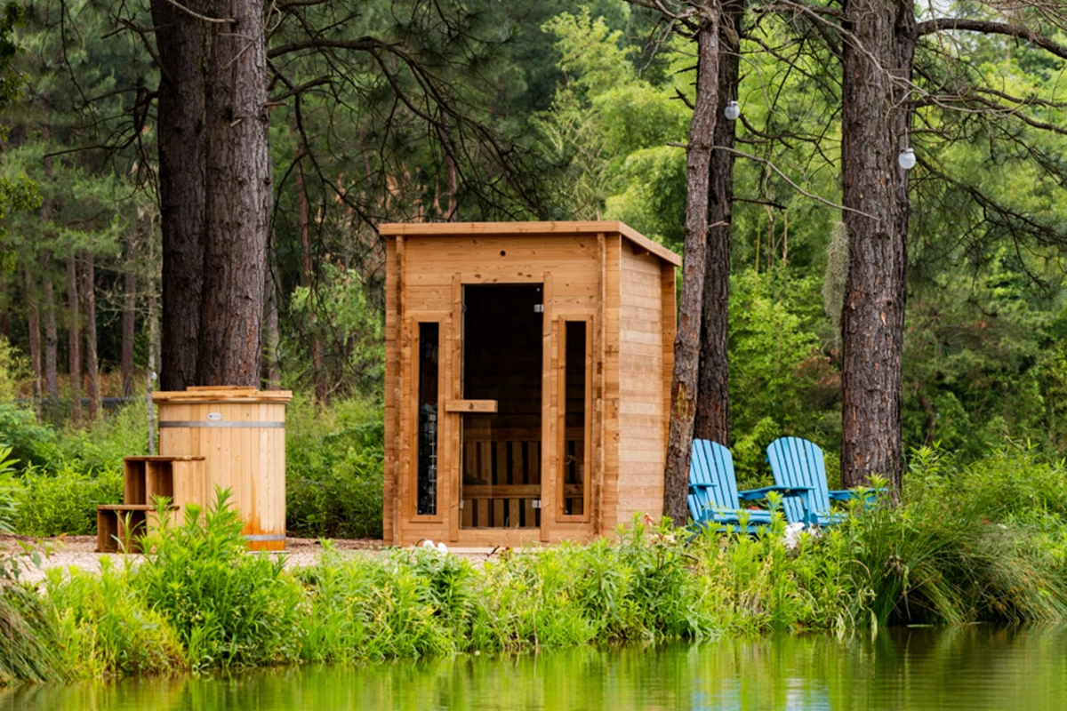 collections-hot-tub-wellness-redwood-1 Wooden sauna hut beside a pond in a forest, with two blue Adirondack chairs nearby outdoorsy setting.