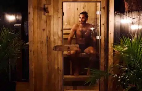 collections-hot-tub-wellness-redwood-4 Man seated inside a small wooden sauna cabin, looking to the side behind a glass door with warm lighting and greenery around.