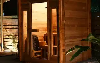 Wooden sauna hut at night with a glass door, warm interior, bench and heater visible inside.