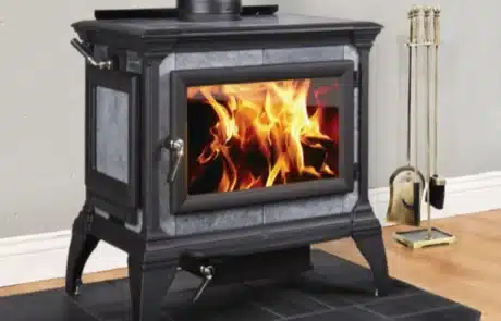 specials-fireplace-stove-1 Black freestanding wood stove with glass door showing orange flames, on a tiled hearth with a fire tool set nearby.
